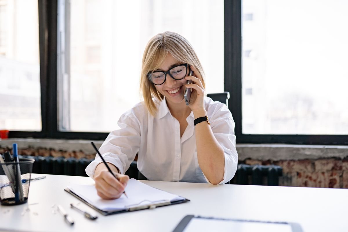 A woman sits at a desk in a modern office, talking on the phone and taking notes